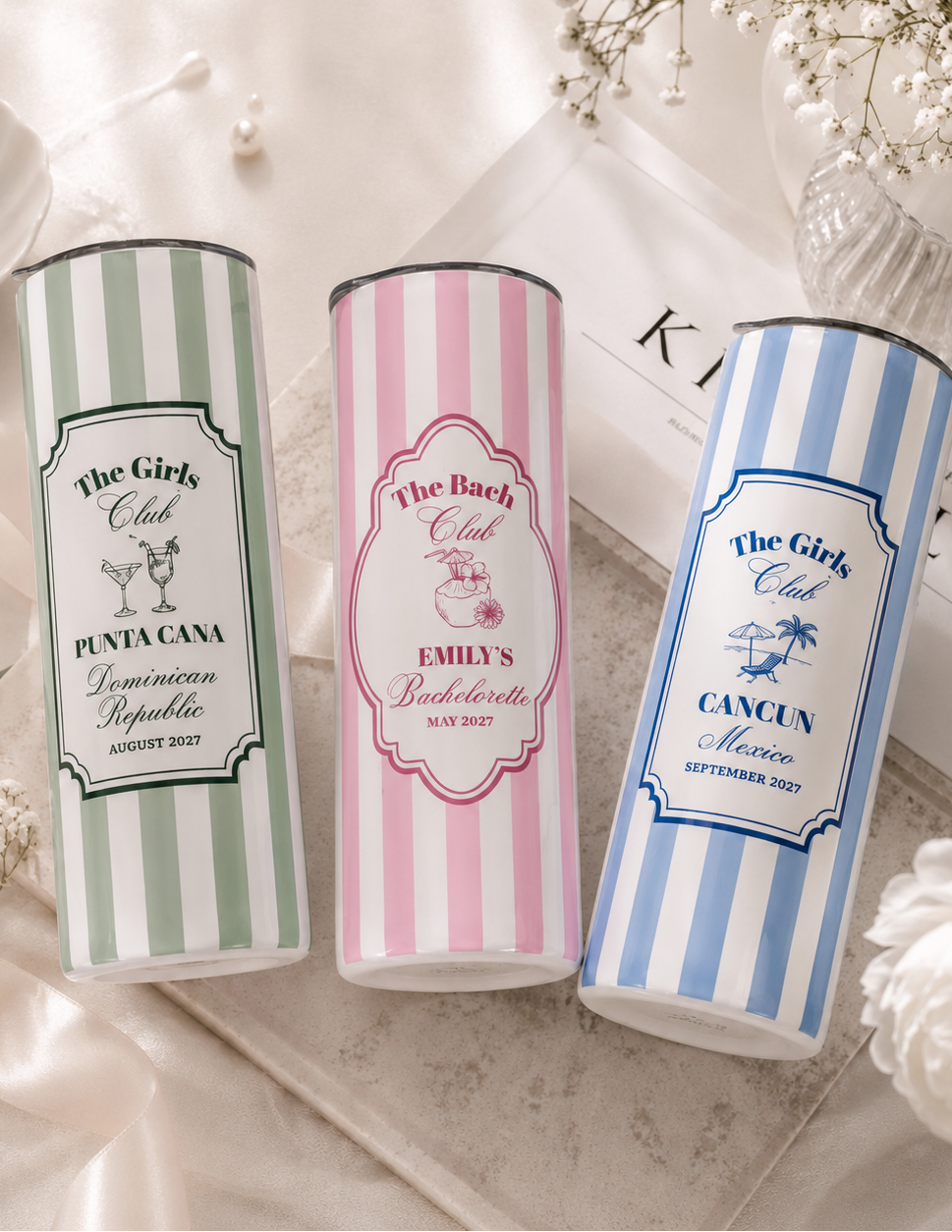 Three personalized cylindrical tumbler cups with vertical stripes are arranged on a light fabric surface with decorative flowers and pearls. The left cup is white and sage green with the text 'The Girls Club' and 'Punta Cana Dominican Republic'; the center cup is white and pink with 'The Bach Club' and 'Emily's Bachelorette'; the right cup is white and blue with 'The Girls Club' and 'Cancun Mexico'.